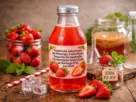 Garrafa de kombucha de morango com rótulo descritivo sobre mesa de madeira, cercada por morangos frescos, cubos de gelo e potes de vidro.
