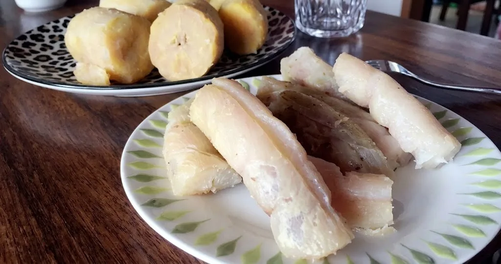 Bobolo: A Fermentação Tradicional da Mandioca na África Central Prato com mandioca cozida ao centro e outro prato ao fundo com bananas cozidas, dispostos sobre mesa de madeira escura com garfo e copo de vidro ao lado. Imagem em formato horizontal, ideal para capas de blog e redes sociais.